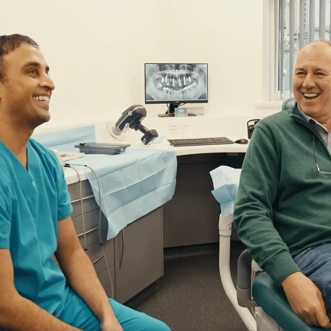 Dentist and male patient smiling together during post-treatment consultation at Infinity Dental Clinic with a dental implant X-ray visible on screen